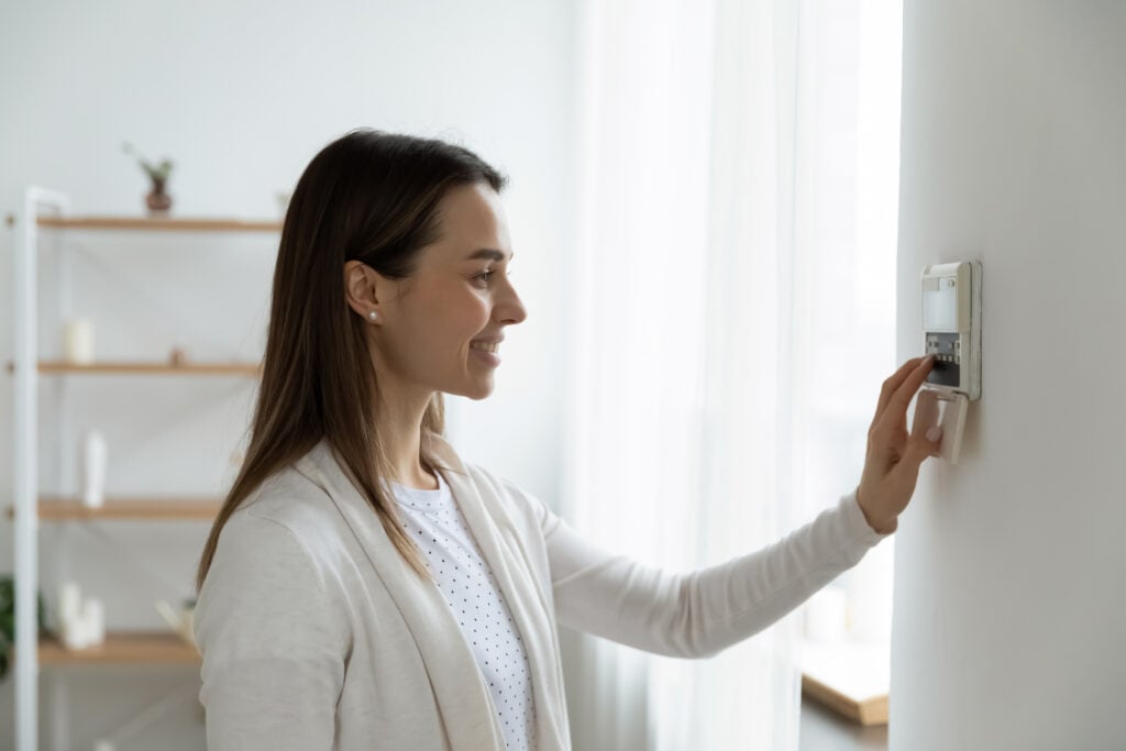 Smiling woman standing near wall-mounted device adjusting degrees in living room set comfortable temperature using thermostat home heating system. Owner of modern smart house, energy saving concept how to install smart thermostat