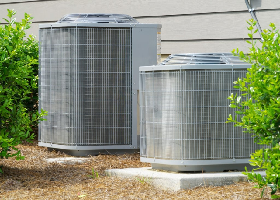 Two gray outdoor air conditioning condenser units surrounded by green shrubbery. The image highlights the primary mechanical system used to manage temperature.