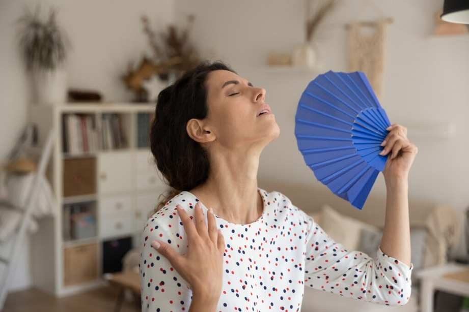 overheated woman fanning herself with a blue hand fan indoors, clearly experiencing discomfort due to high indoor heat and humidity.