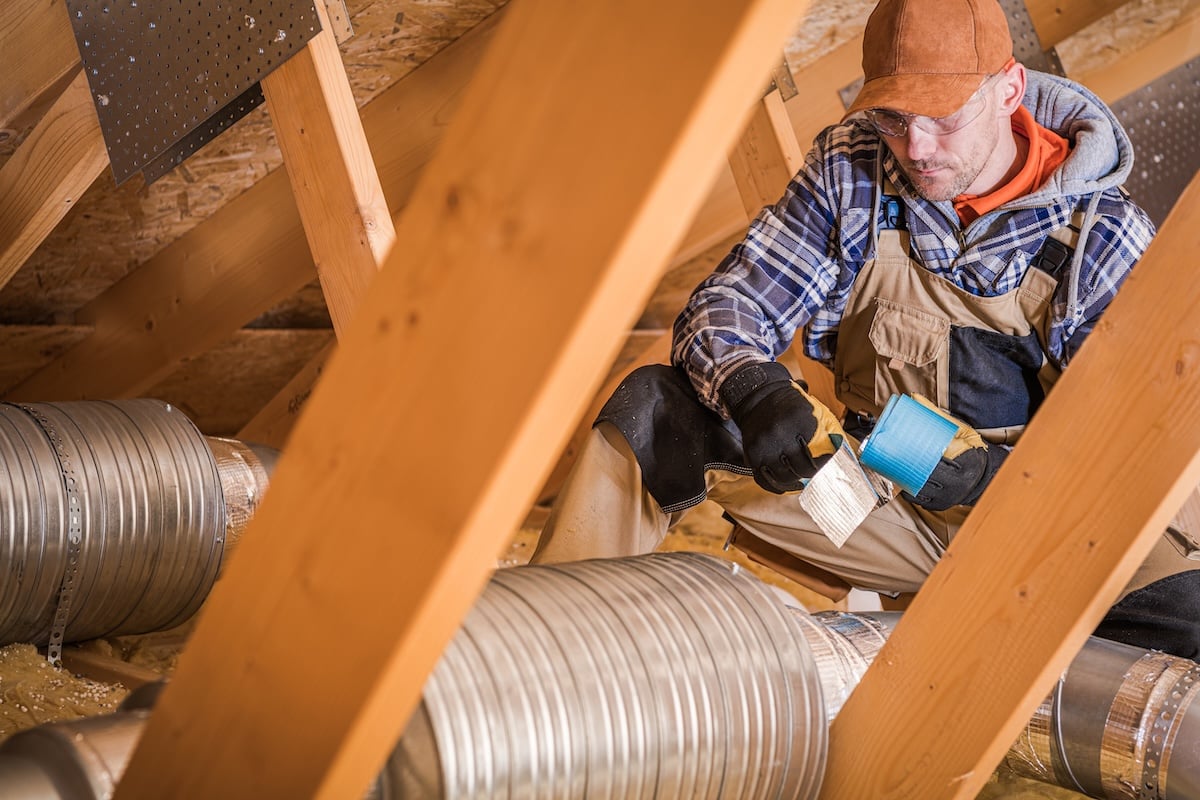 Construction Worker Wrapping HVAC Air Duct With Silver And Blue Foil Tape In Attic Of Residential Building.