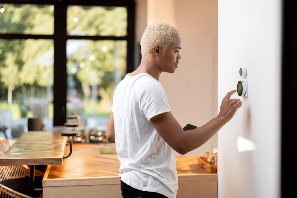 recommended thermostat settings for summer and winter Latin man choosing temperature on thermostat. Young focused guy pushing button on smart home system. Concept of modern domestic lifestyle. Interior of kitchen in modern apartment.