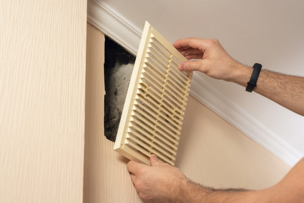 A man installs a plastic ventilation grid into a hole in a wall. mold in air ducts
