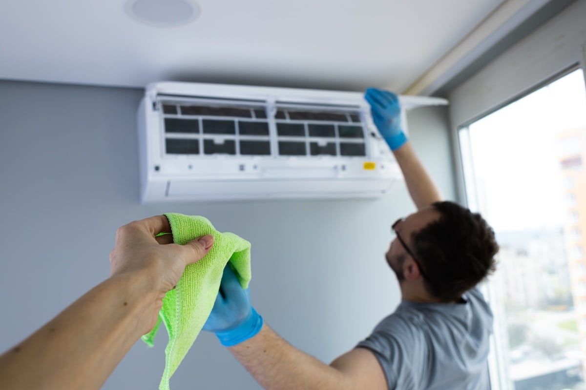 An individual is cleaning an air conditioning unit while another person hands a green cloth, emphasizing the importance of regular maintenance for efficiency and air quality.
