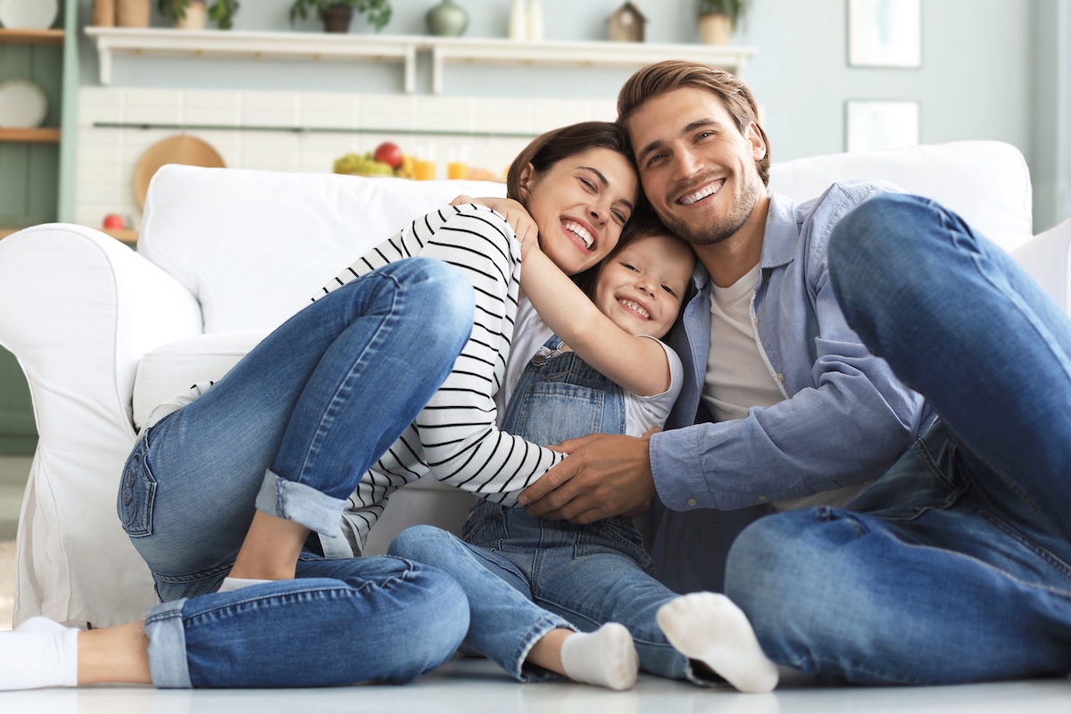 Young Caucasian family with small daughter pose relax on floor in living room, smiling little girl kid hug embrace parents, show love and gratitude, rest at home together