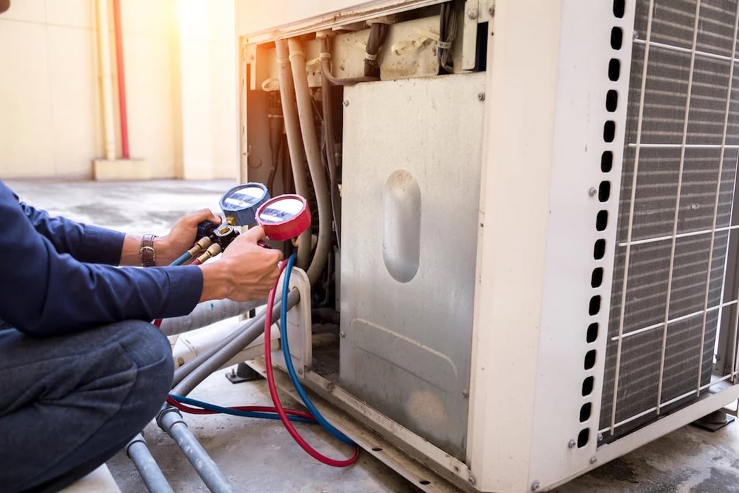 A professional HVAC technician kneels beside an outdoor air conditioning condenser unit, attaching a manifold gauge set to the copper refrigerant lines to test the system's pressure levels.