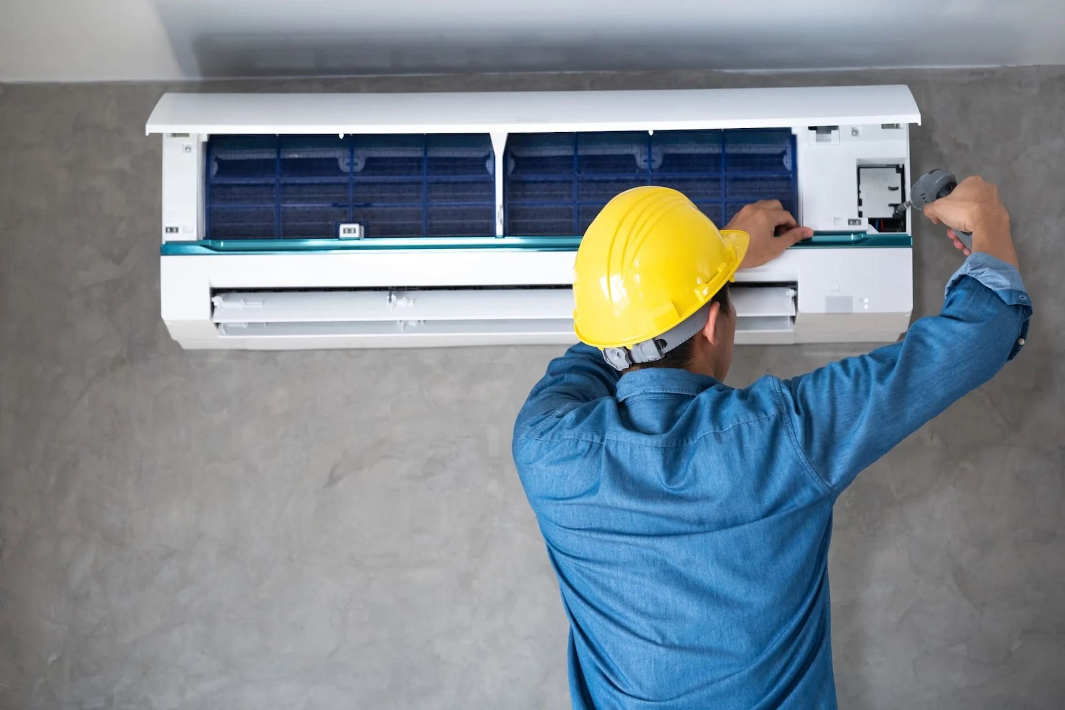 A skilled heating and cooling specialist wearing a yellow safety helmet carefully services the internal components of a white ductless mini-split air conditioner mounted against a textured gray wall.