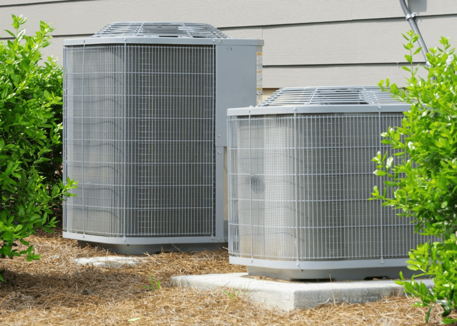 Two large exterior air conditioning condenser units rest securely on concrete pads alongside a home with tan siding.