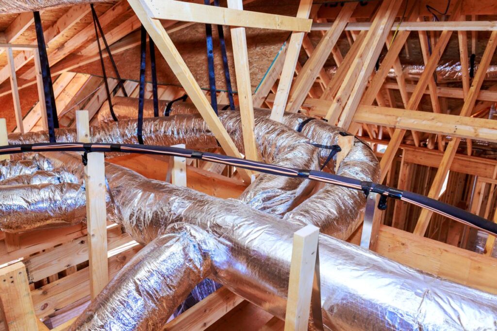 Workers install metal ductwork among wooden beams in home attic setting during renovation process.